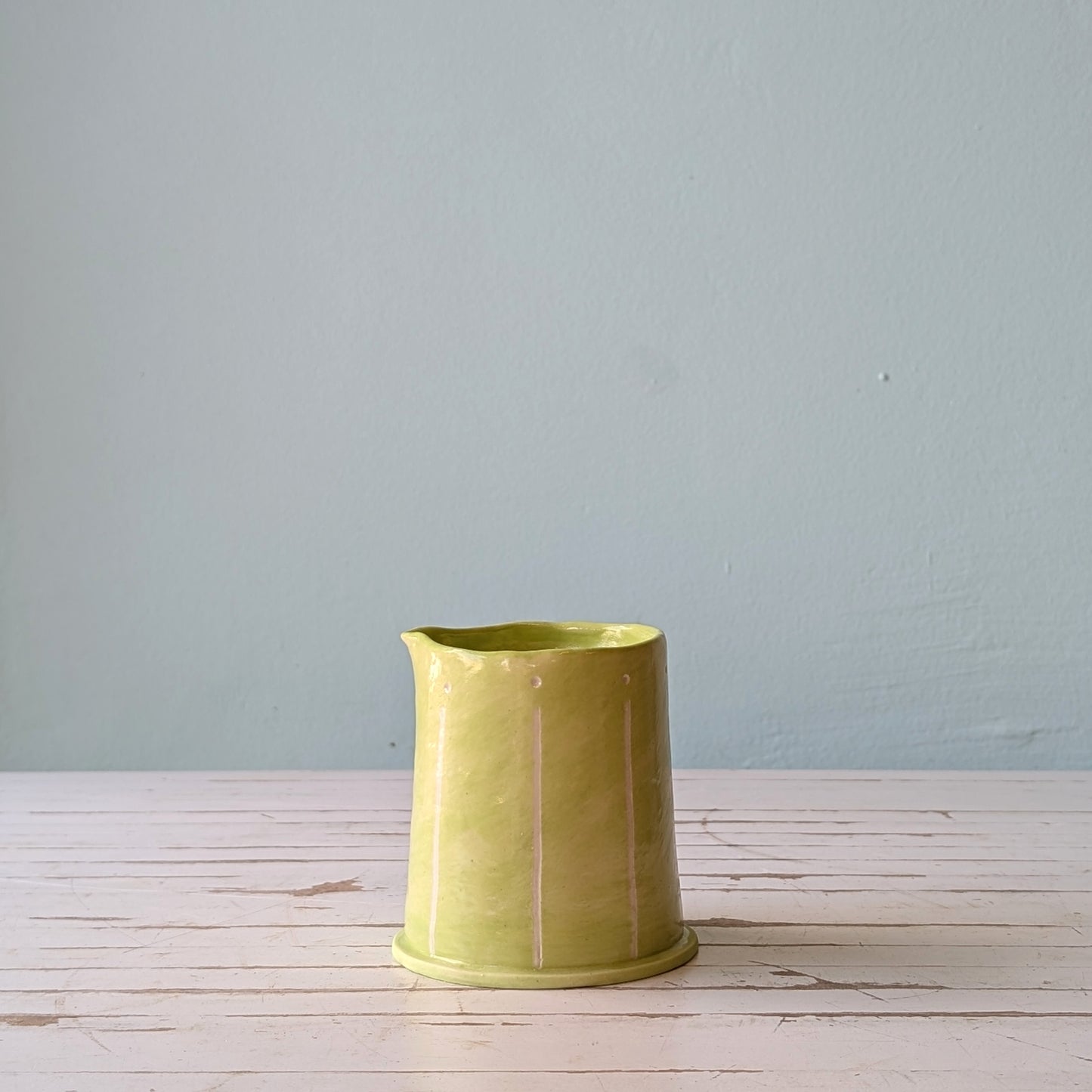 Front view of a light green ceramic pourer on a white wooden surface against a blue background.