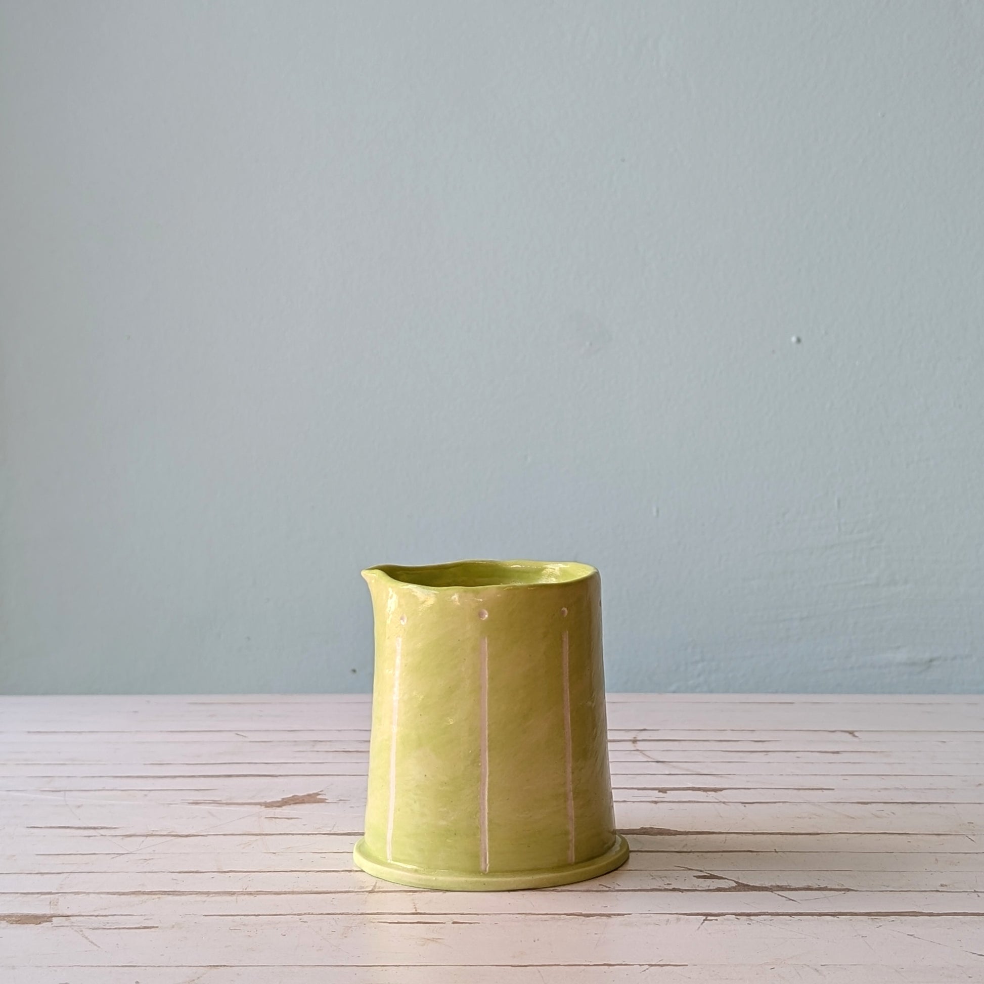 Front view of a light green ceramic pourer on a white wooden surface against a blue background.