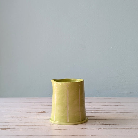 Front view of a light green ceramic pourer on a white wooden surface against a blue background.
