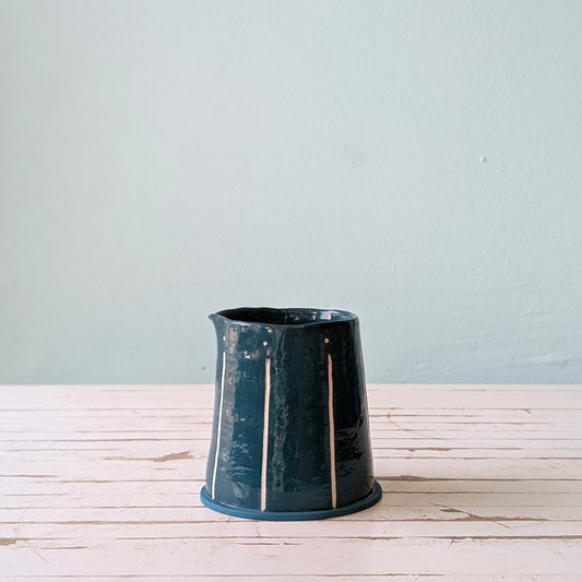 Front view of a blue/green ceramic pourer on a white wooden surface against a blue background.