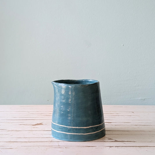 Front view of a blue ceramic pourer on a white wooden surface against a blue background.