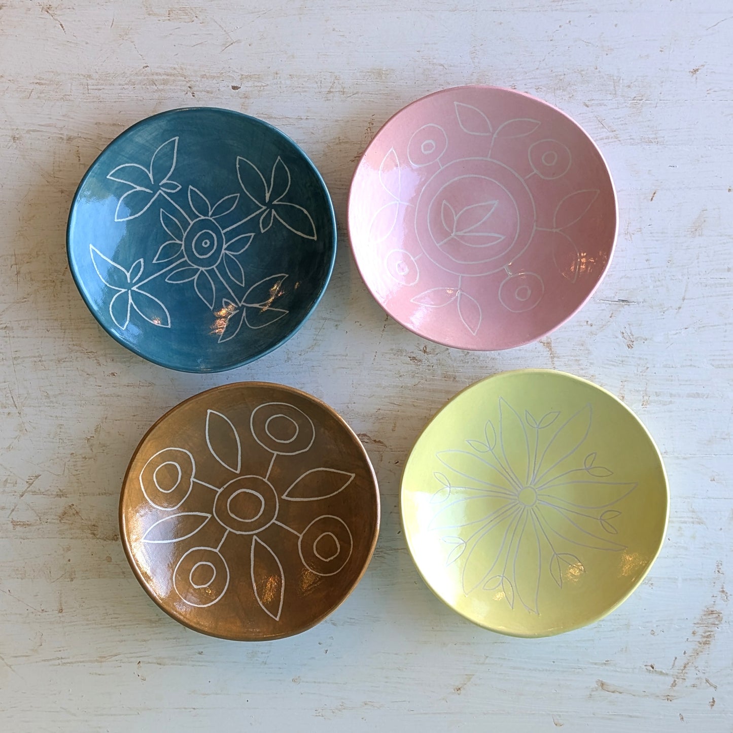 Four small ceramic bowls arranged on a light distressed wooden surface. The bowls are teal blue, pink, bronze-brown, and pale yellow, each decorated with a white hand-drawn style floral and circular pattern.