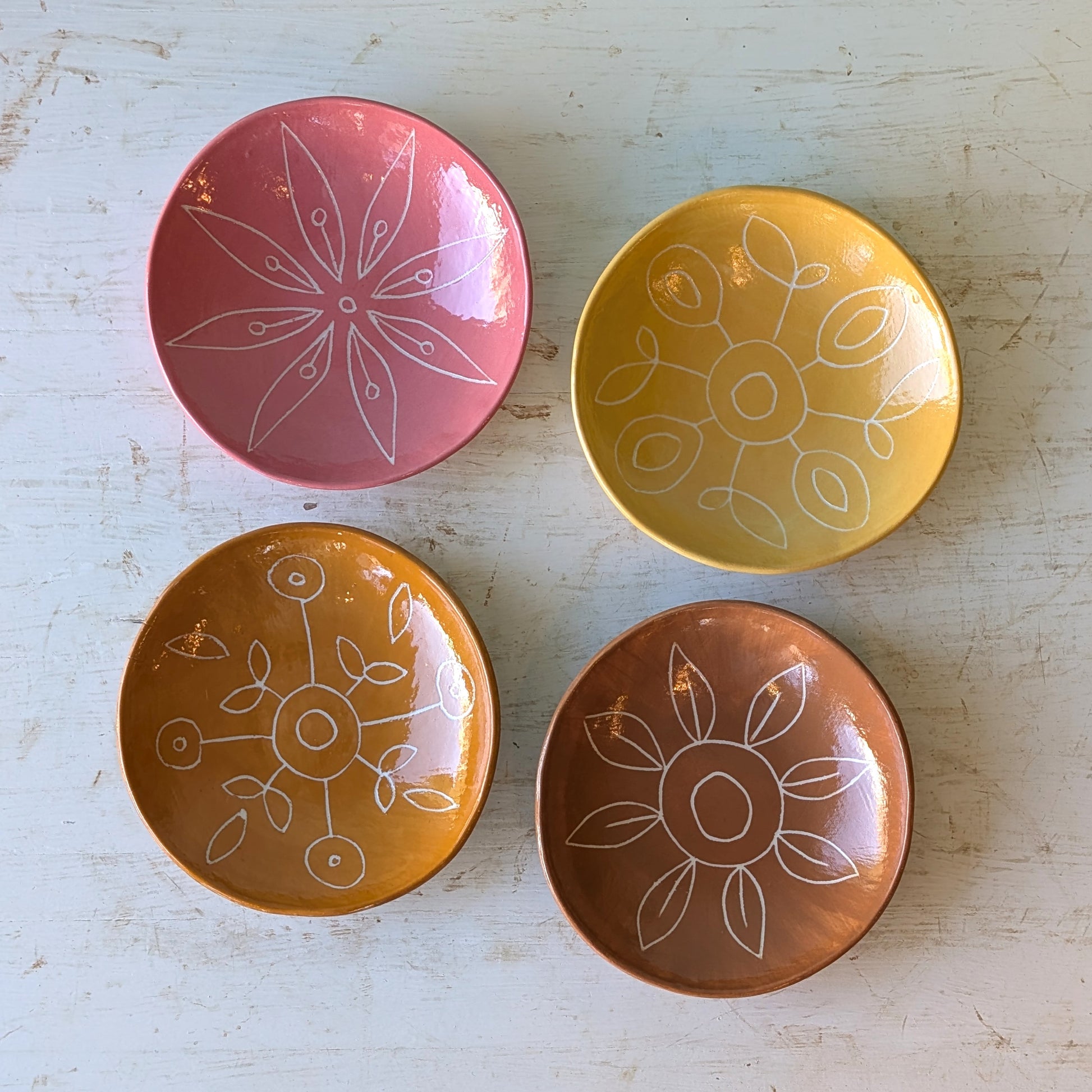 Four small round ceramic plates in pink, yellow, orange, and brown, each decorated with a white hand-drawn floral or geometric pattern, arranged on a distressed white wooden surface.