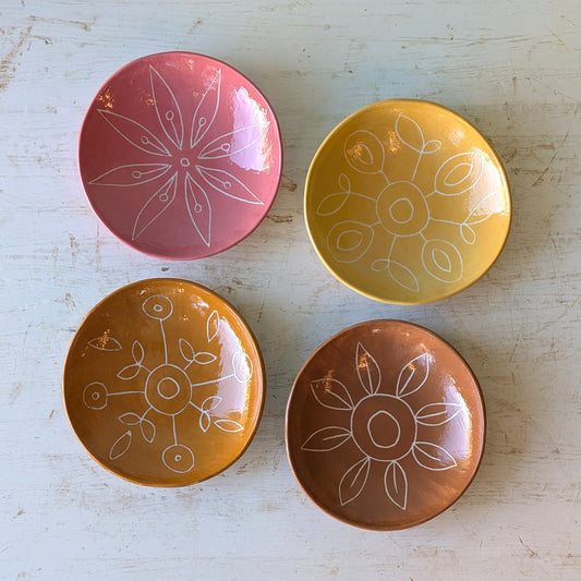 Four small round ceramic plates in pink, yellow, orange, and brown, each decorated with a white hand-drawn floral or geometric pattern, arranged on a distressed white wooden surface.