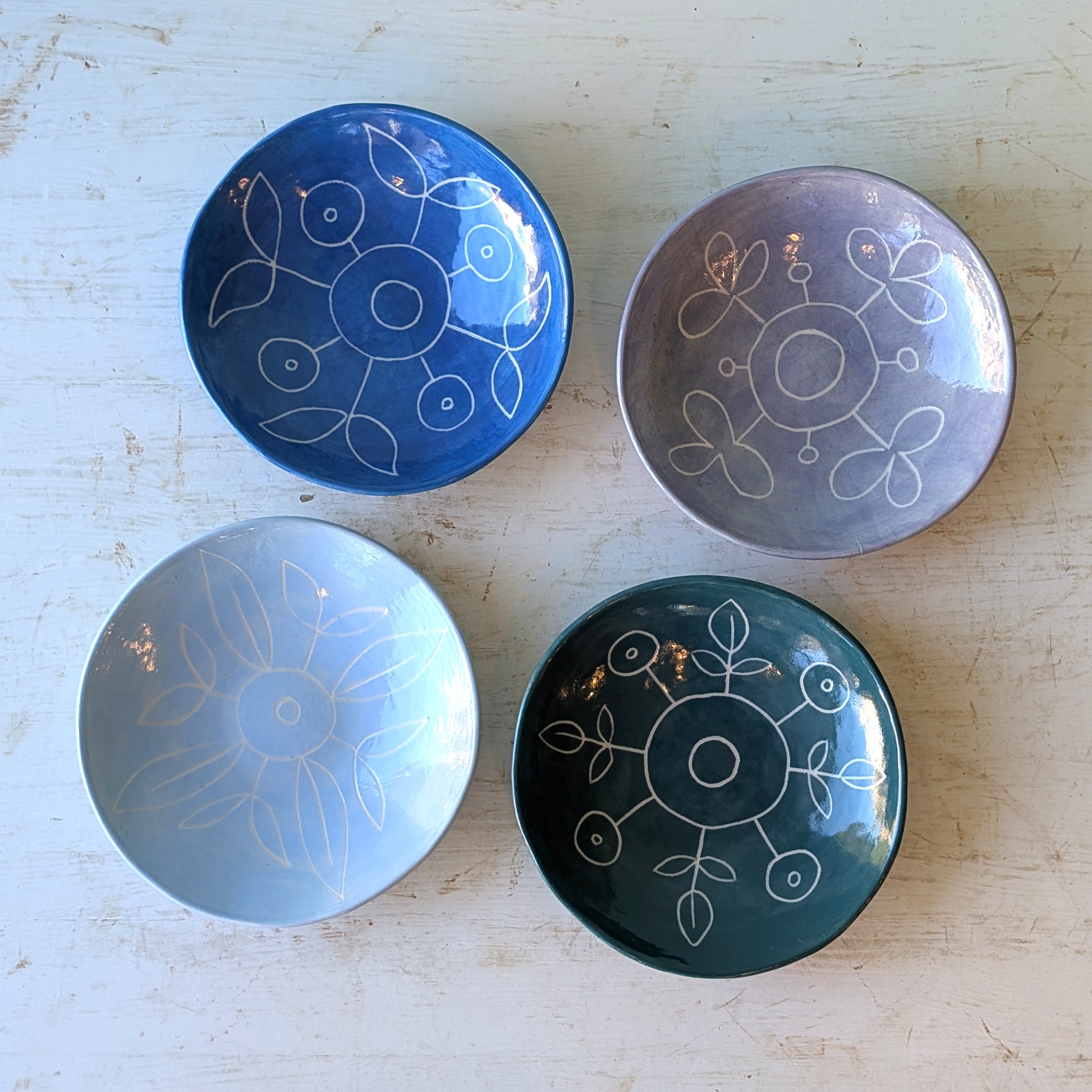 Four small ceramic bowls with hand-painted floral designs in blue, purple, light blue, and dark green, arranged on a distressed white wooden surface.