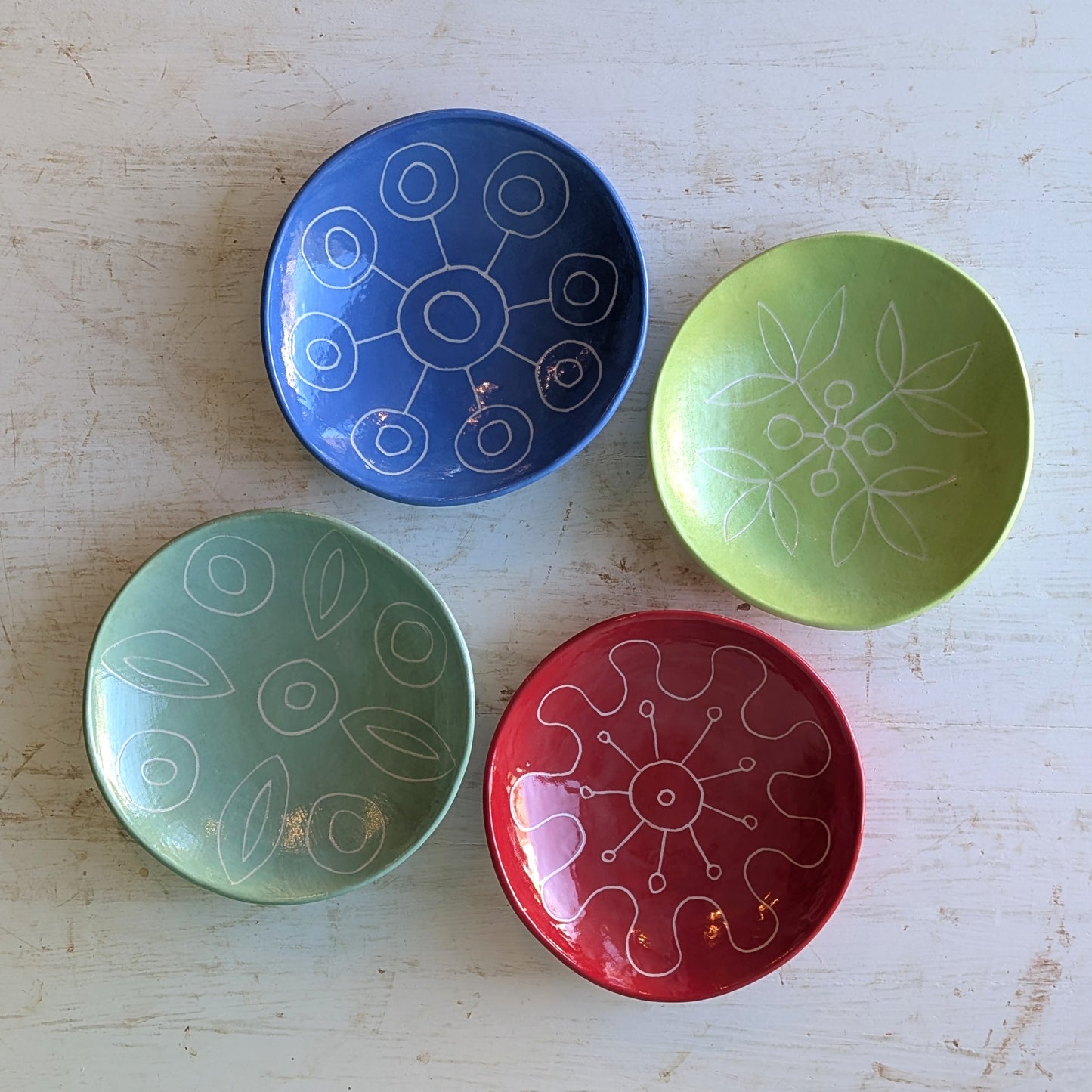 Four small ceramic bowls in blue, green, and red, each decorated with white hand-drawn circular and floral patterns, arranged on a light wood surface.
