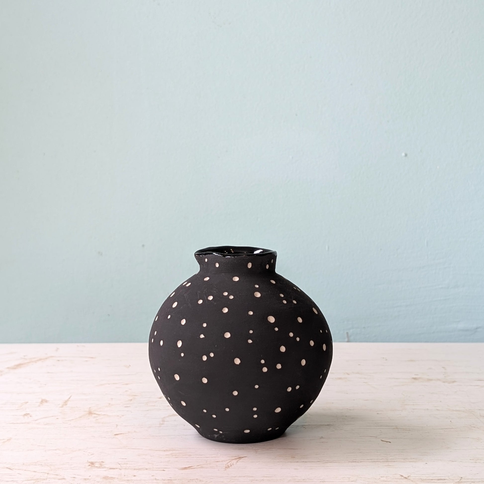 Black vase with white polka dots on a light wooden surface with a light blue background