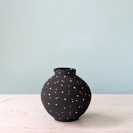 Black vase with white polka dots on a light wooden surface with a light blue background