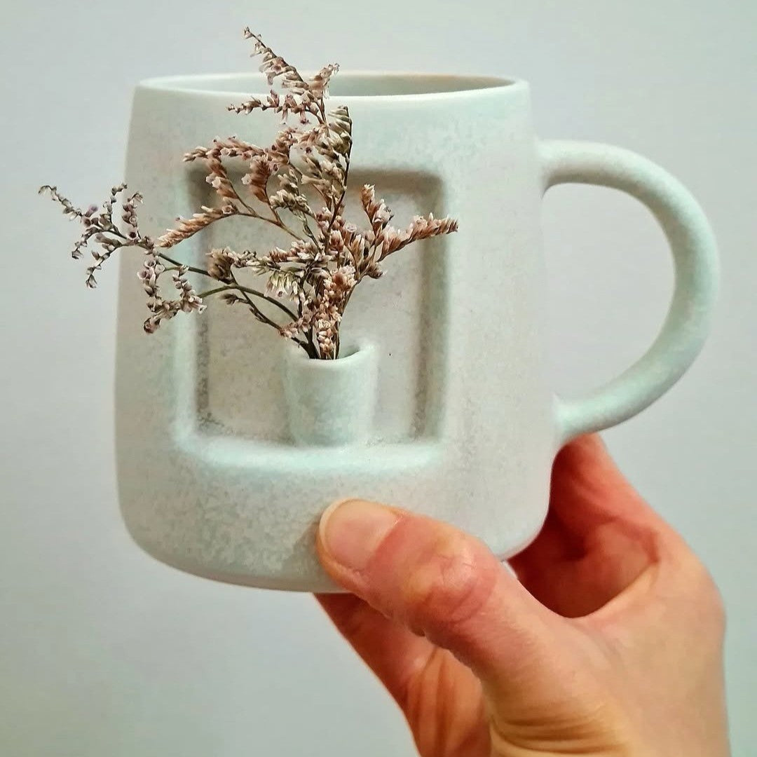 Light blue mug with a unique design holding a sprig of flowers, held by a hand against a plain background
