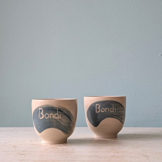 Two white cups with a blue splash of glaze and the word "Bondi" written on them, on a white wooden table against a light blue wall.