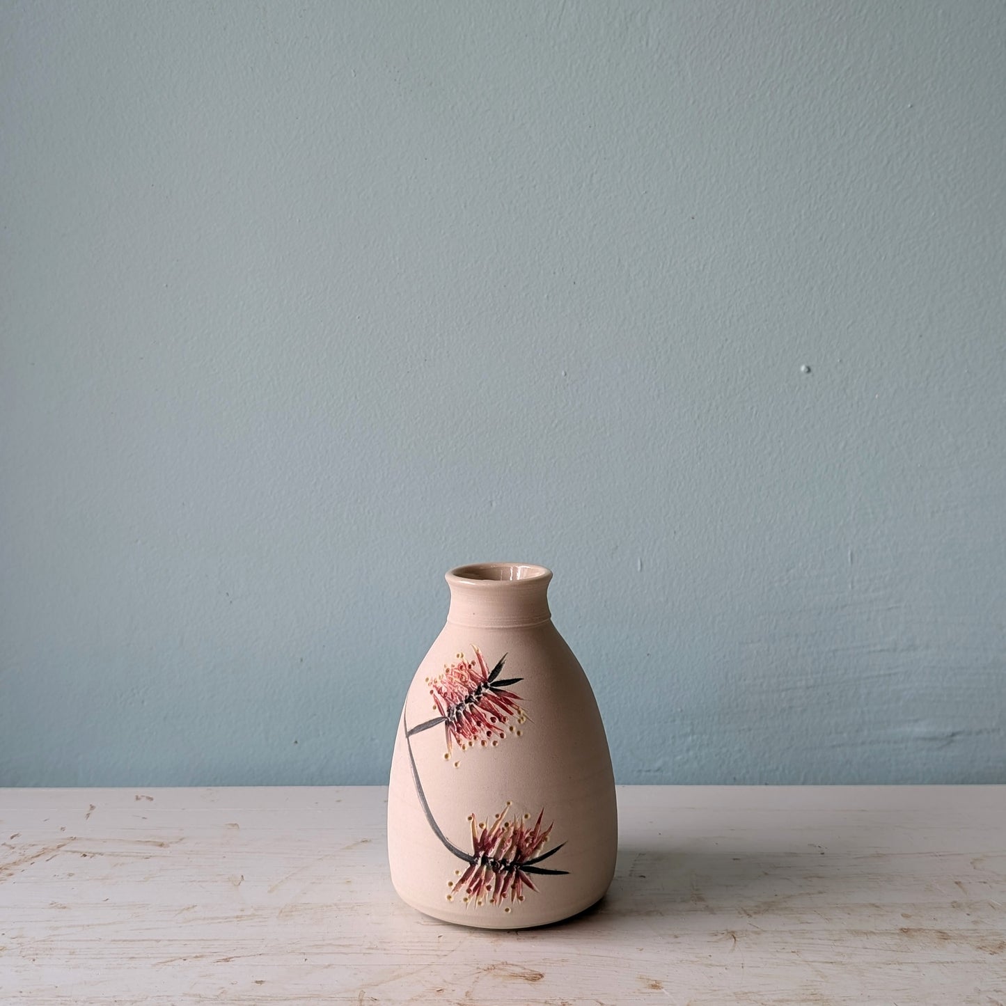 Ceramic vase with floral bottlebrush pattern on a white table against a blue wall.