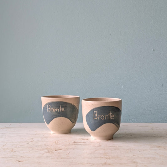 Two white cups with a blue splash of glaze and the word "Bronte" written on them, on a white wooden table against a light blue wall.