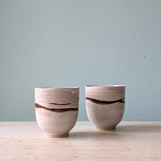 Two handmade ceramic cups with a textured beige glaze and a dark brown horizontal crack pattern, displayed on a light wooden surface against a muted blue wall.