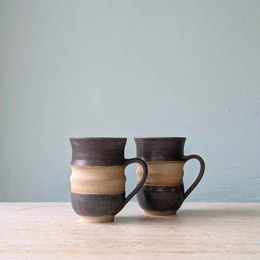 Two charcoal and beige ceramic mugs with handles and textured surface, on a white wooden table against a light blue wall.