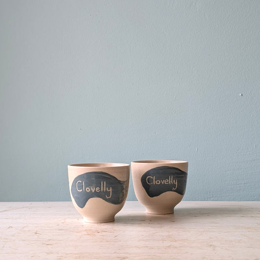 Two white cups with a blue splash of glaze and the word "Clovelly" written on them, on a white wooden table against a light blue wall.
