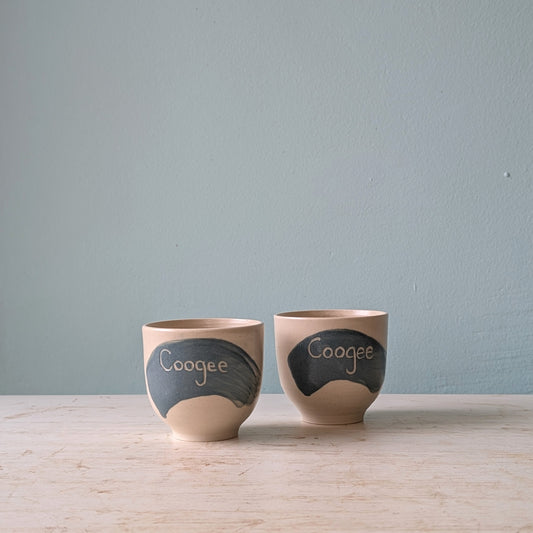 Two white cups with a blue splash of glaze and the word "Coogee" written on them, on a white wooden table against a light blue wall.