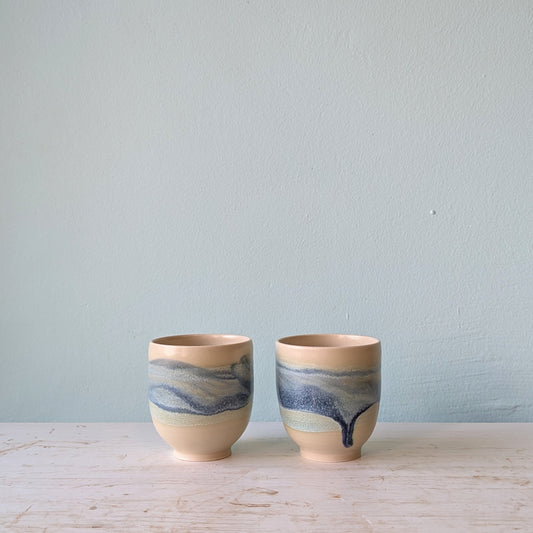 Two beige and blue ceramic cups on a white table against a blue wall.