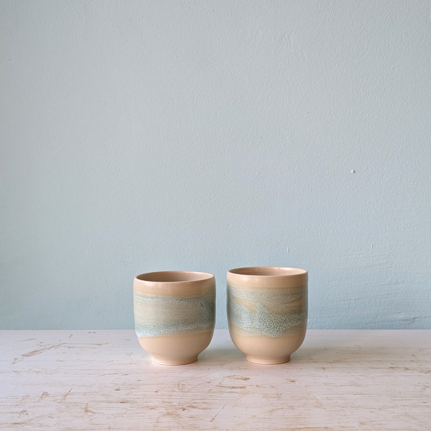 Two beige and light green cups on a white table against a blue wall.