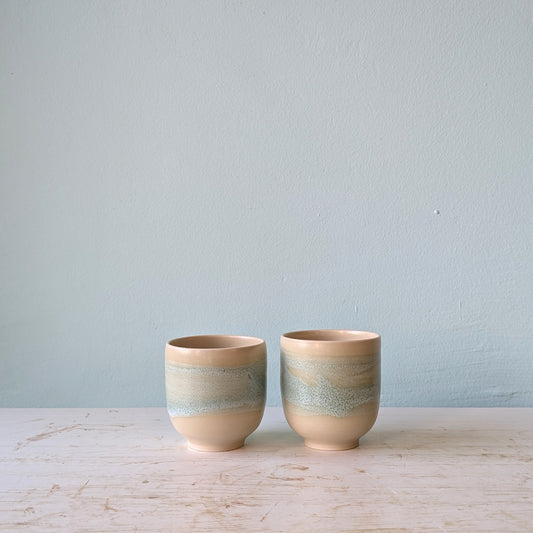Two beige and light green cups on a white table against a blue wall.