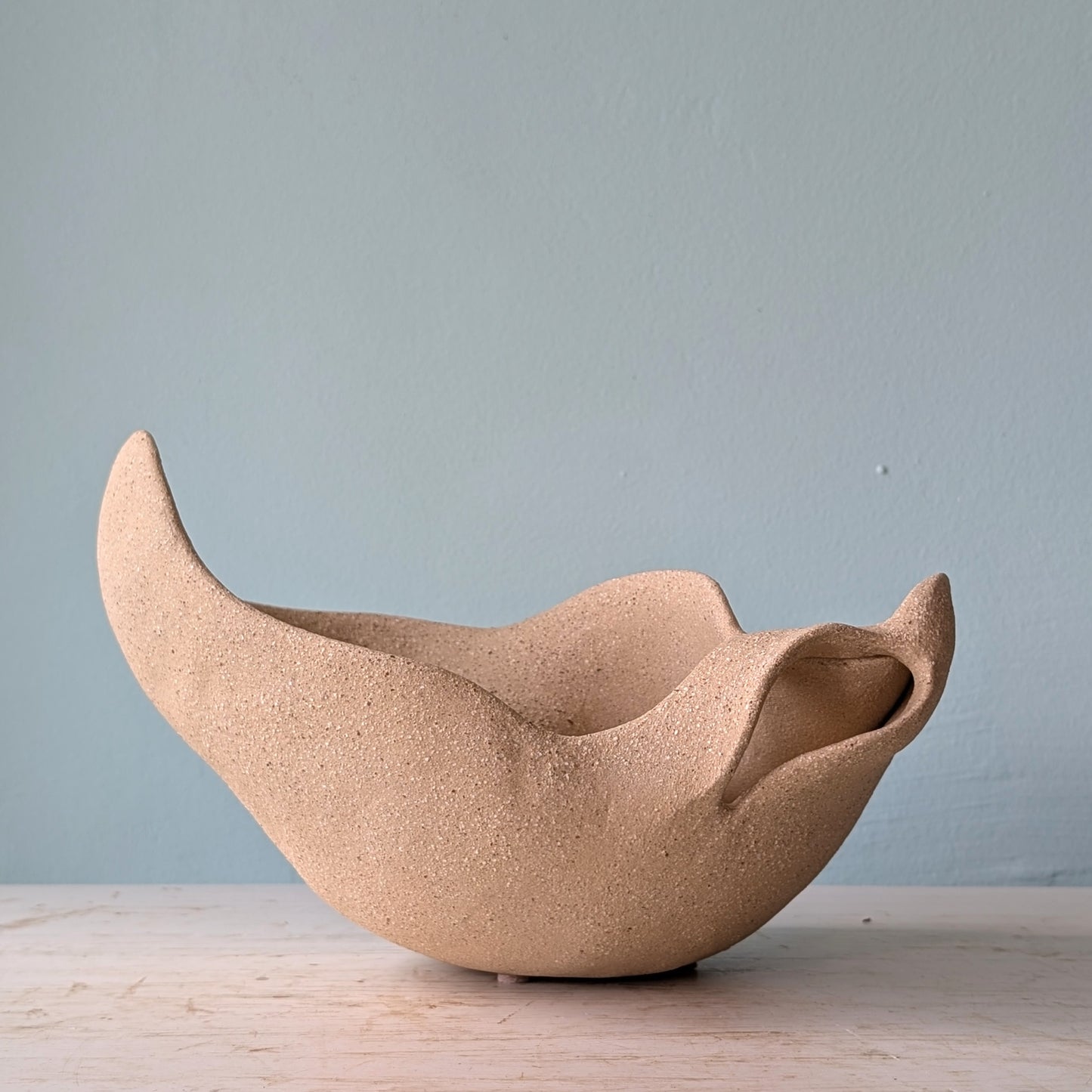 Sculptural bowl made of beige matte ceramic on a white table against a blue wall.