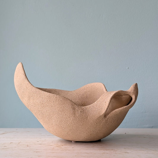Sculptural bowl made of beige matte ceramic on a white table against a blue wall.