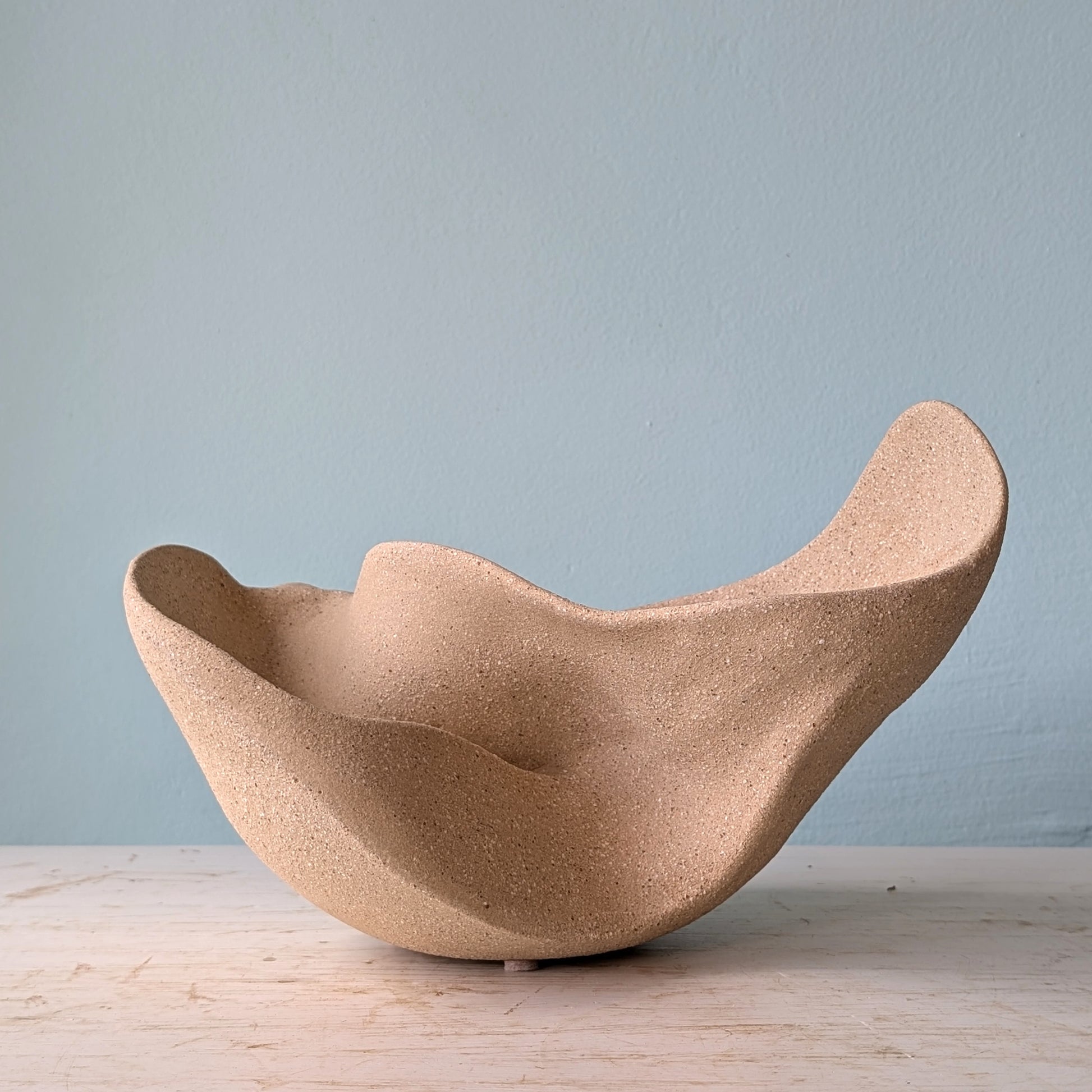 Sculptural bowl made of beige matte ceramic on a white table against a blue wall.
