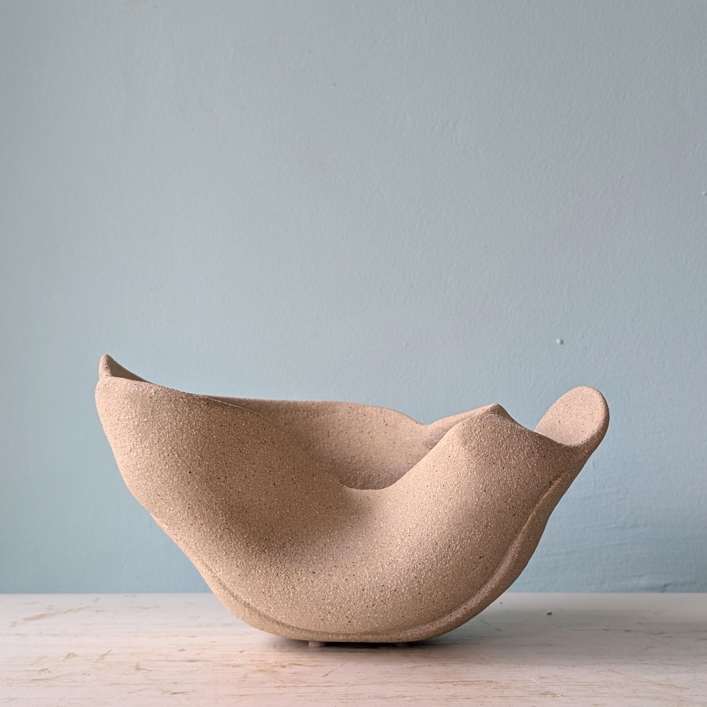 Sculptural bowl made of beige matte ceramic on a white table against a blue wall.