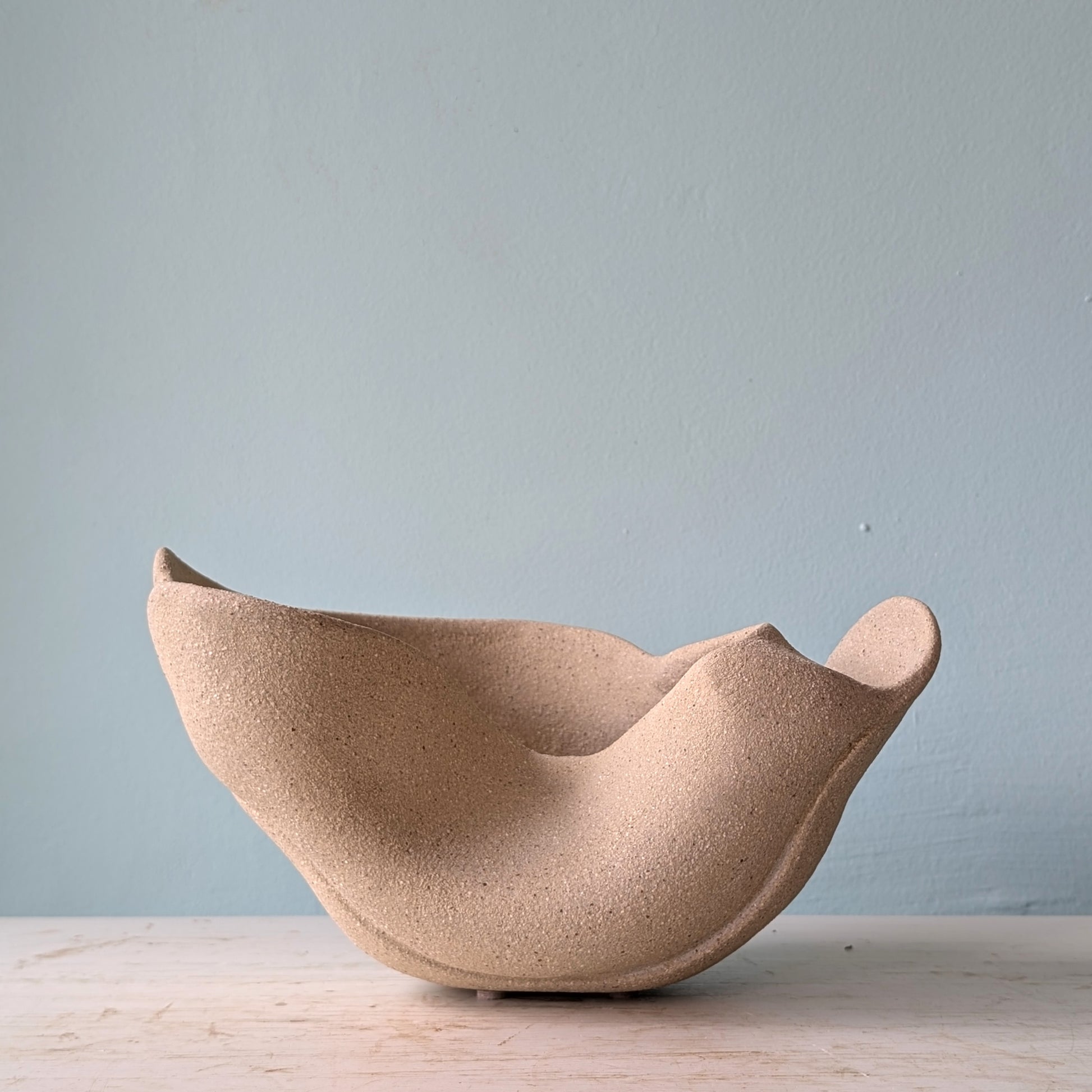 Sculptural bowl made of beige matte ceramic on a white table against a blue wall.