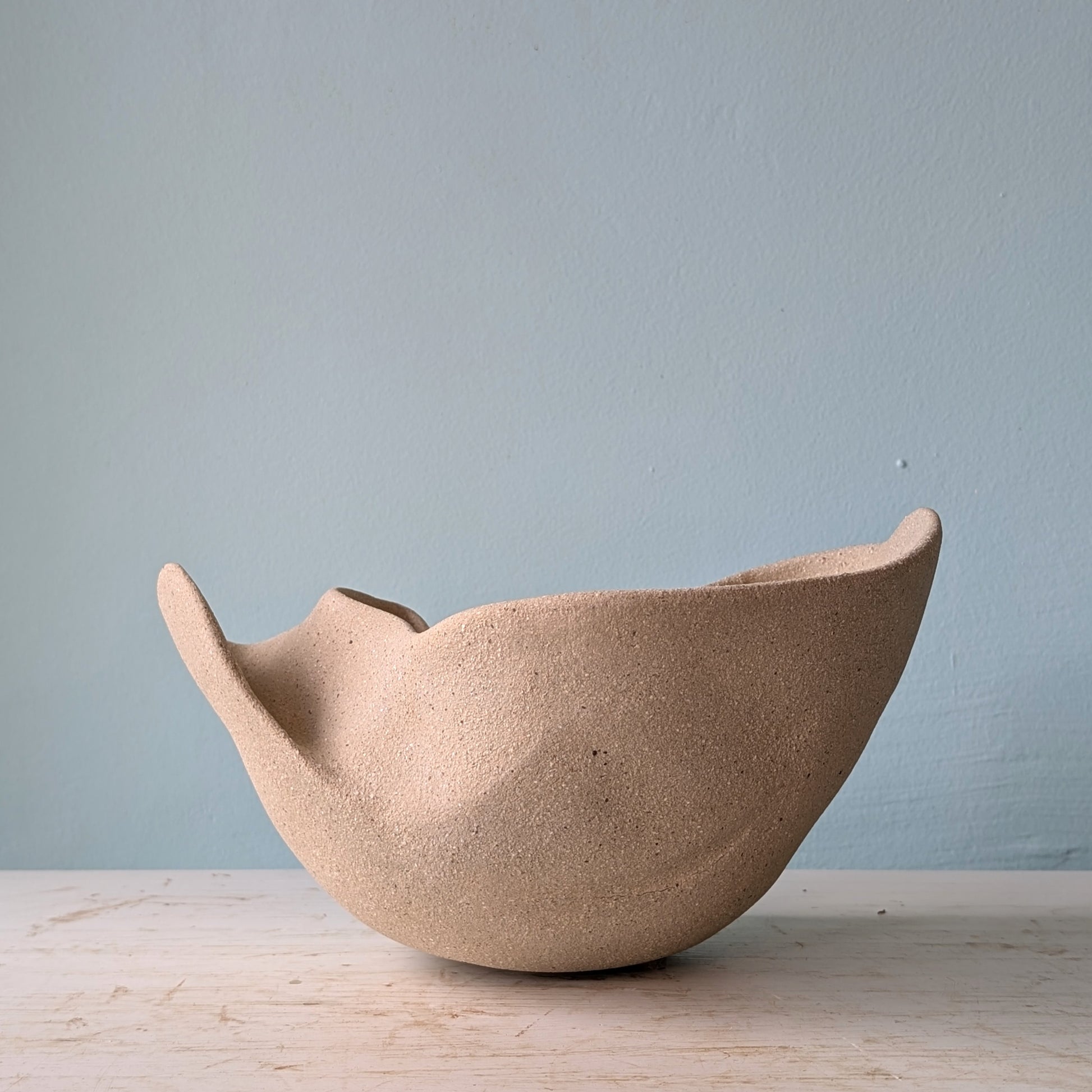 Sculptural bowl made of beige matte ceramic on a white table against a blue wall.