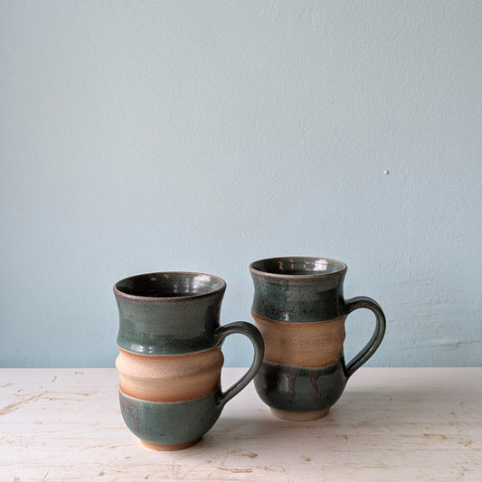 Two forest green ceramic mugs on a white table against a blue wall.
