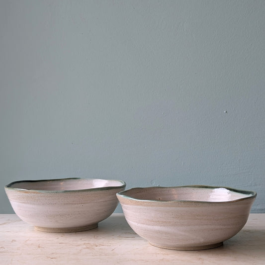 Two handmade ceramic bowls with a textured beige finish and a dark green rim, displayed on a light wooden surface against a muted blue wall.