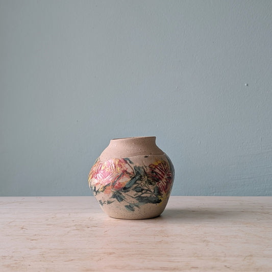 A small round ceramic vase with a natural beige stoneware body and a hand-painted floral design in red, green, and yellow, sitting on a light wooden surface against a muted blue wall.