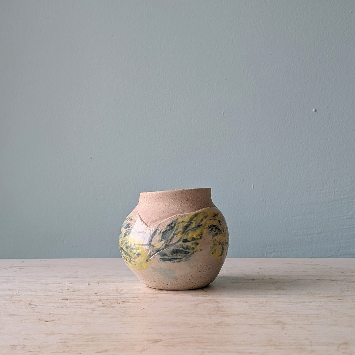 Small ceramic vase with hand painted floral design in yellow and green, on a white wooden table against a light blue wall.