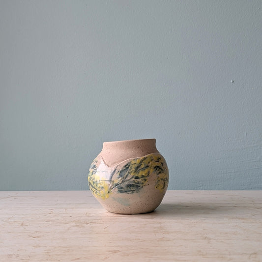 Small ceramic vase with hand painted floral design in yellow and green, on a white wooden table against a light blue wall.