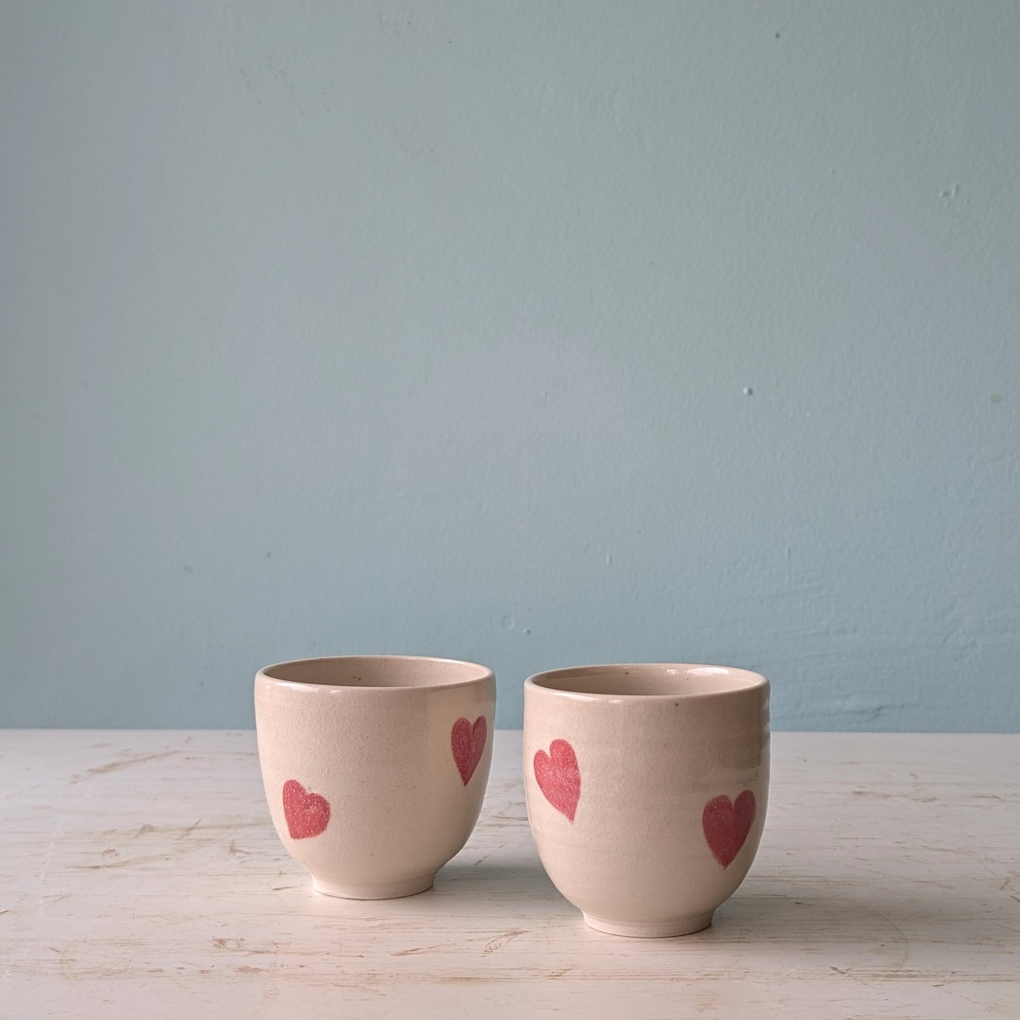 Beige ceramic cups with pink heart design on a white wooden table against a blue wall