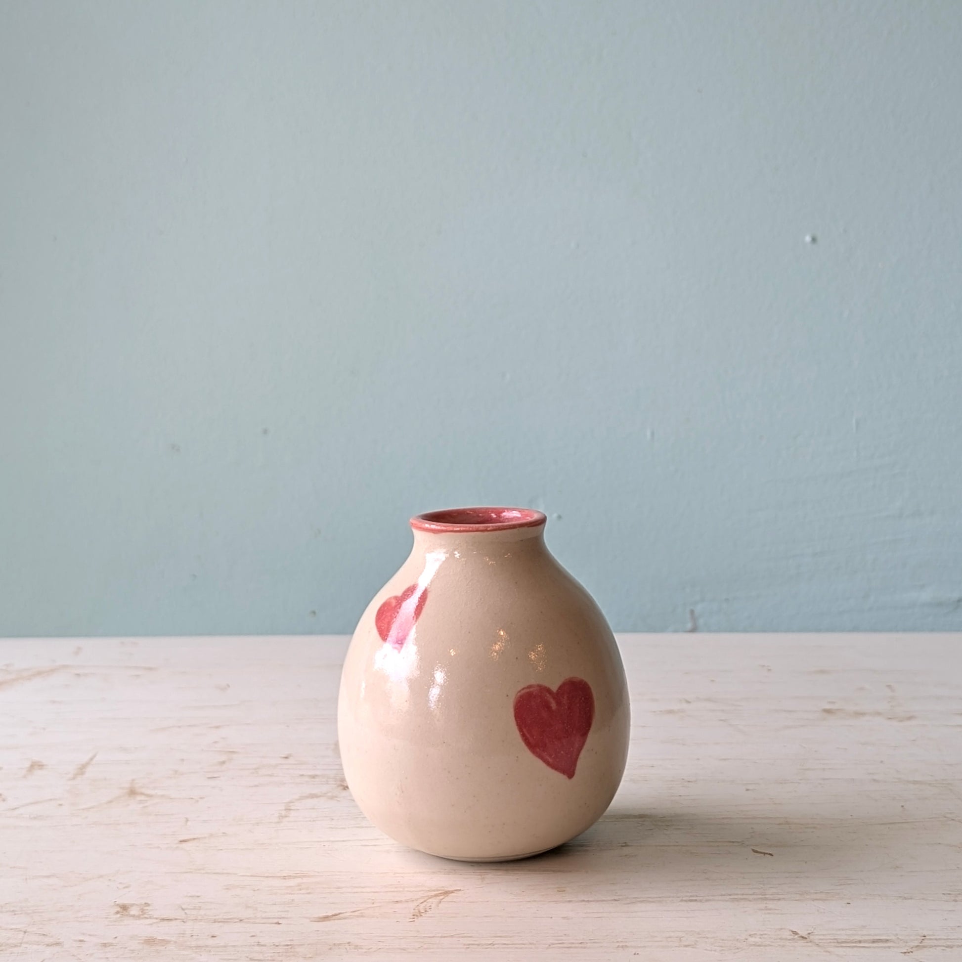 Small ceramic vase with pink hearts on a light wooden surface and blue background