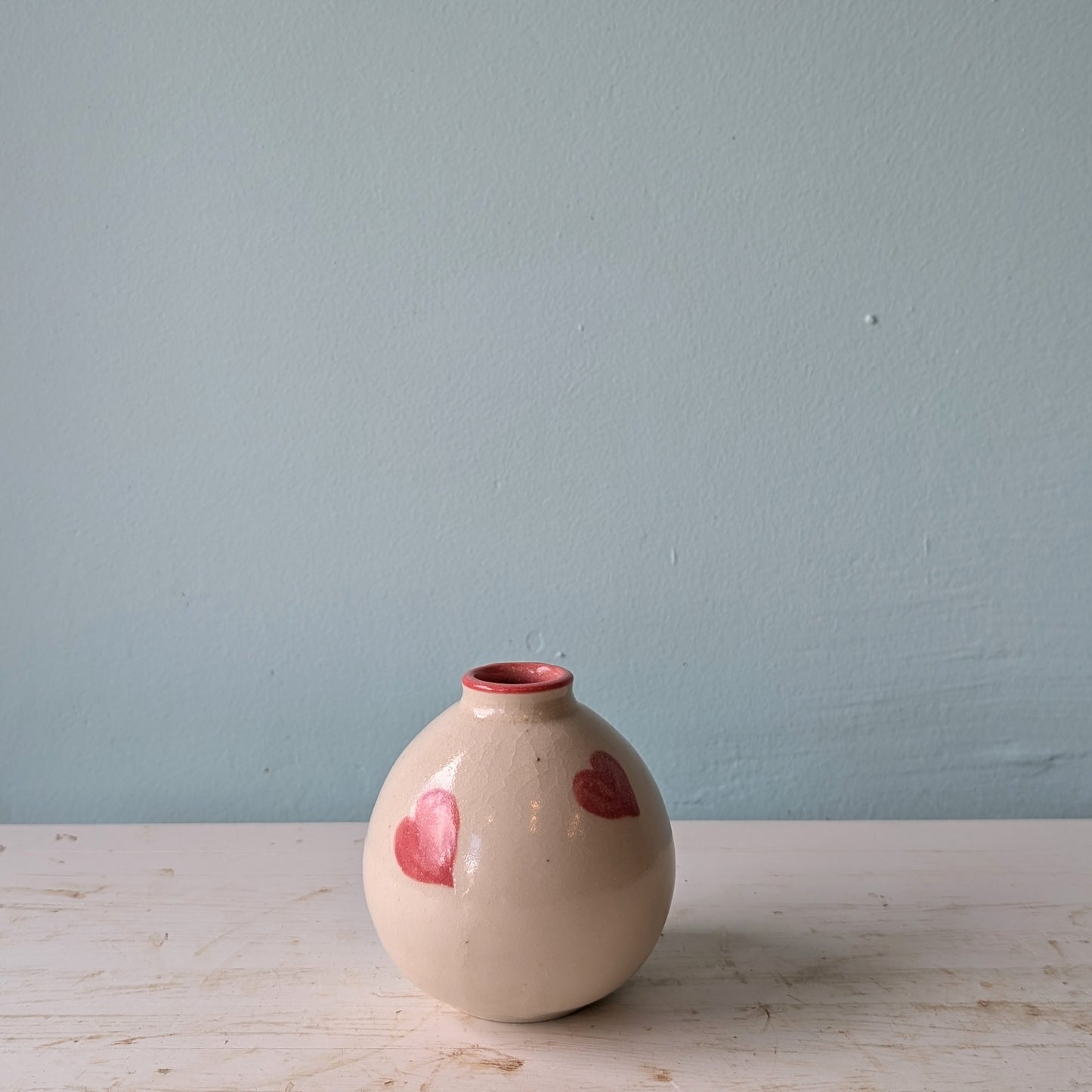 Beige ceramic vase with pink hearts on a white table against a blue wall.