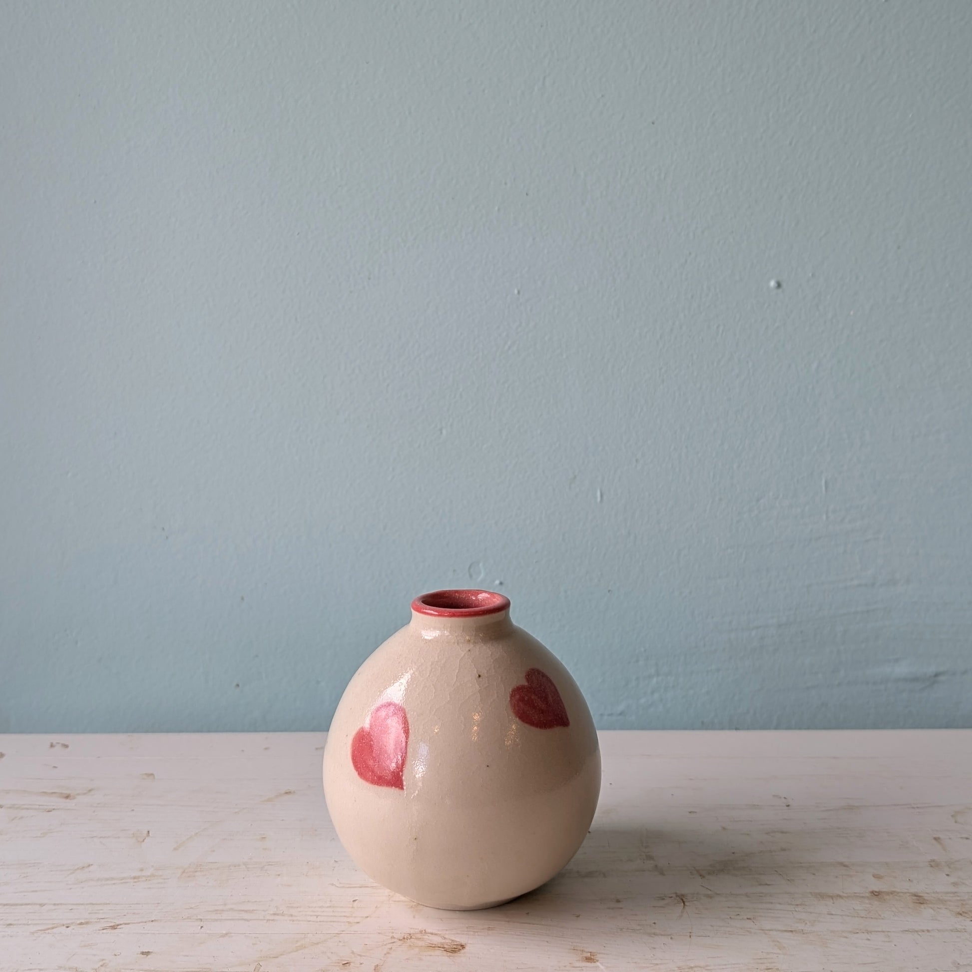 Beige ceramic vase with pink hearts on a white table against a blue wall.