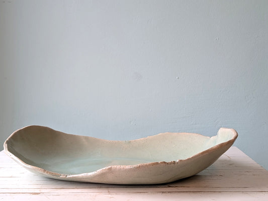 Front view of a freeform ceramic serving platter n light green, on a white wooden table with a blue background.