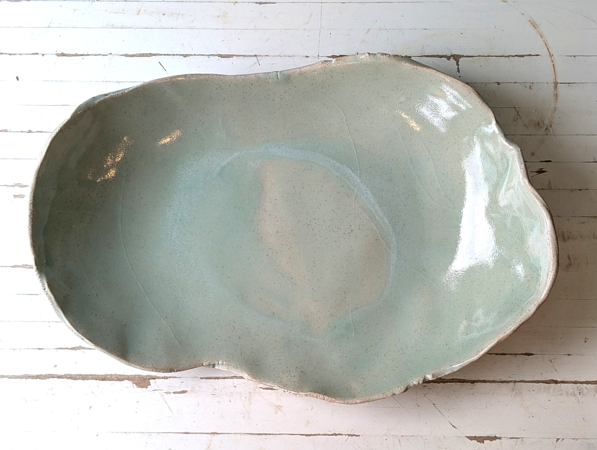 Top view of a freeform ceramic serving platter in light green, on a white wooden table.
