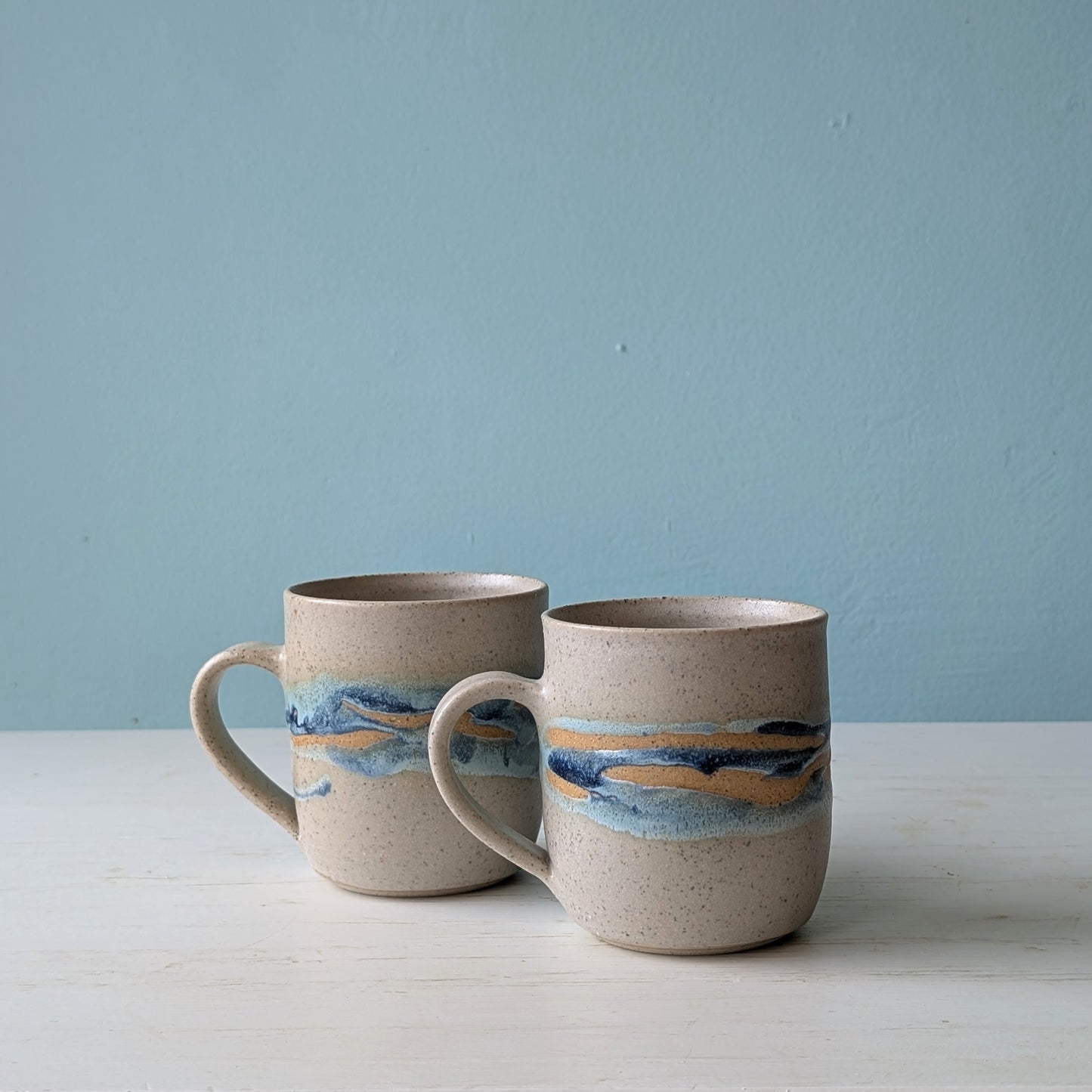 Two ceramic mugs with blue/green glaze pattern on white table against a blue wall