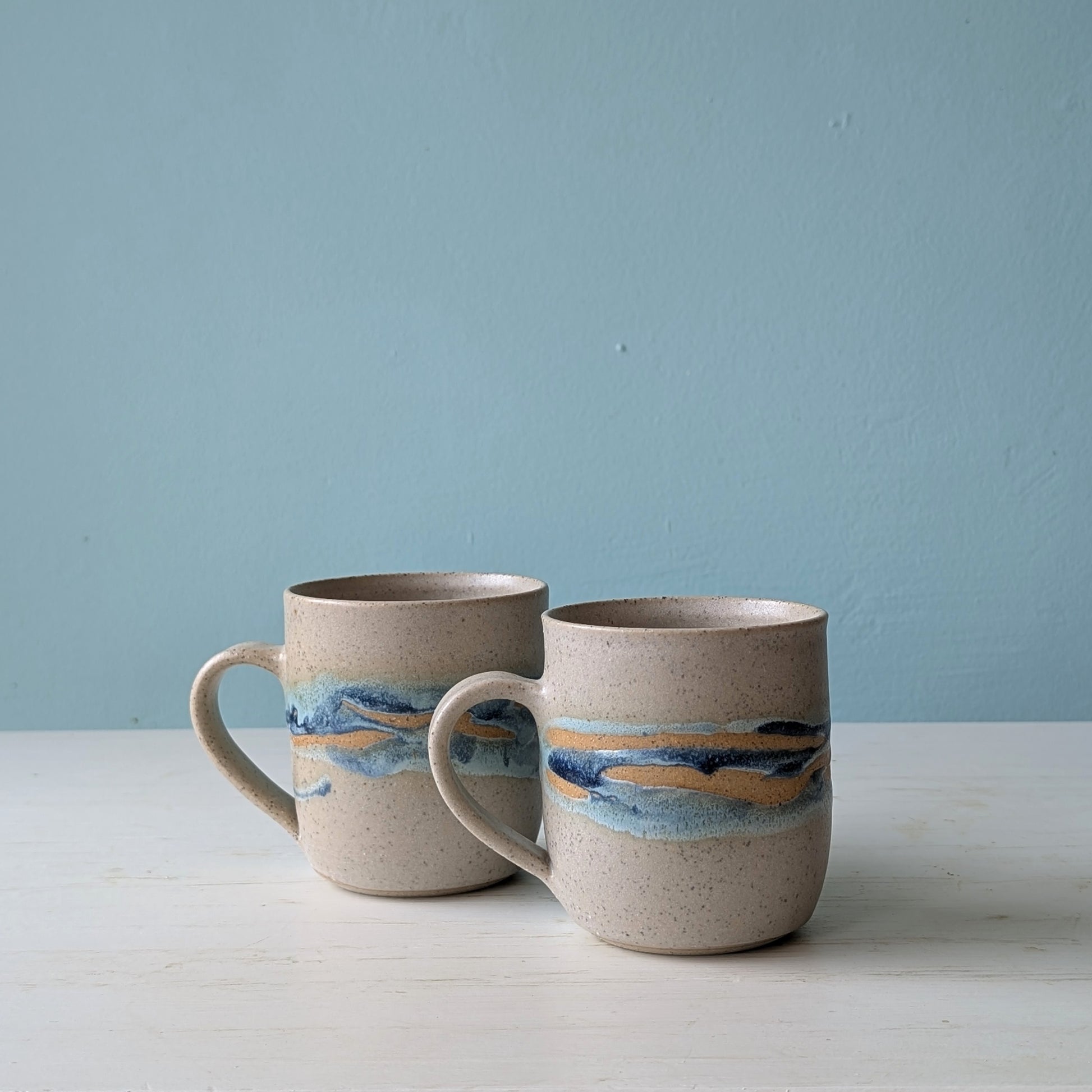 Two ceramic mugs with blue/green glaze pattern on white table against a blue wall