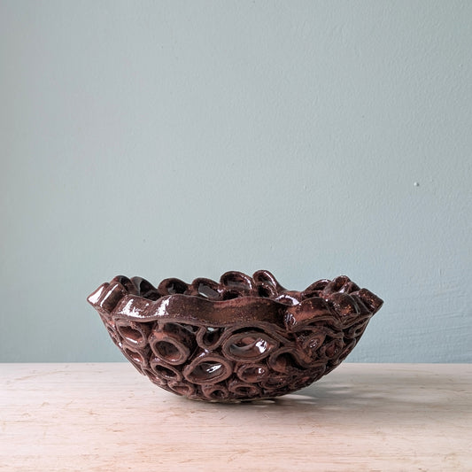 A glossy dark brown ceramic bowl with an intricate, openwork circular pattern, resting on a light wood surface against a muted blue wall.