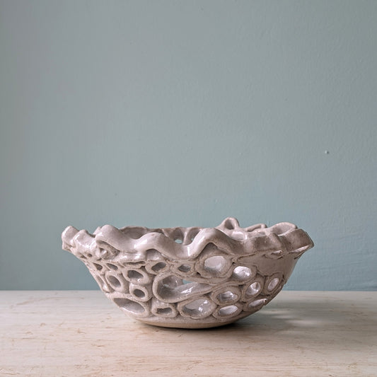 Textured ceramic bowl with lace like pattern, in beige/white, on a white wooden table against a light blue wall.