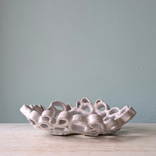 An organic-shaped ceramic bowl with a highly textured, porous, coral-like structure and a matte off-white glaze, displayed on a light wood surface against a muted sage blue wall.