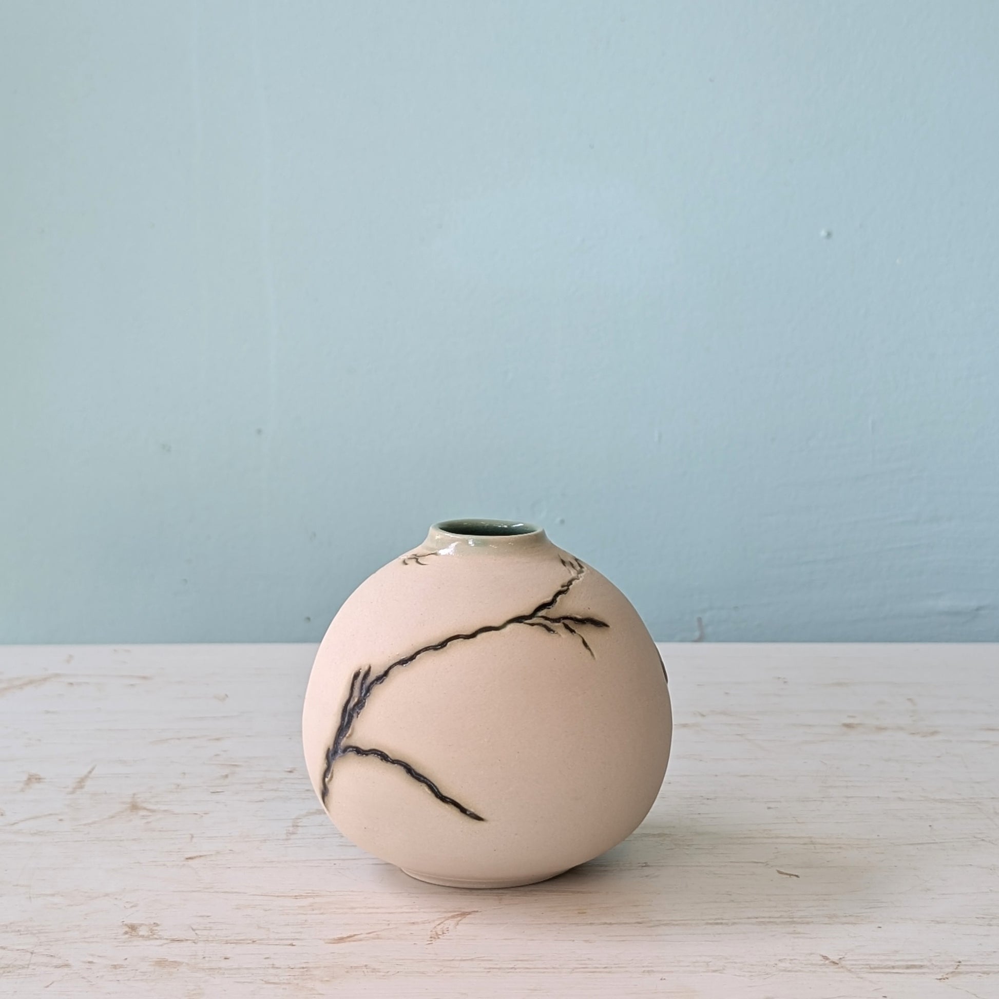 Beige ceramic vase with green seagrass design on a light wooden surface with a light blue wall background