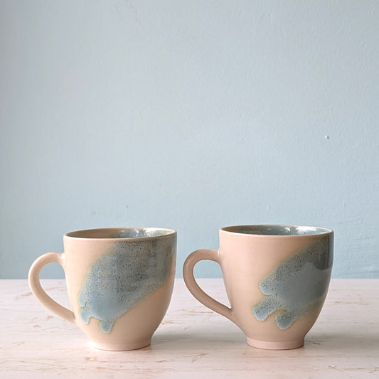 Two ceramic mugs with a speckled design on a light wooden surface and light blue background