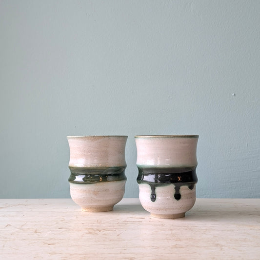 Two handmade ceramic cups with a textured beige and white glaze, featuring a dark green band and a glossy black drip accent, displayed on a light wooden surface against a muted blue wall.