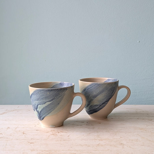 Two ceramic mugs with handles in beige with a blue swirl, on a white tables against a light blue wall.