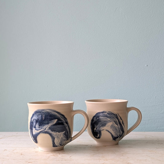 Two ceramic mugs with handles in beige with a dark blue wave, on a white tables against a light blue wall.